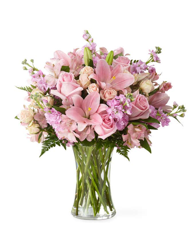 A bouquet of pink lilies, roses, stock and alstroemeria, with fresh floral greens in a tall cylinder vase